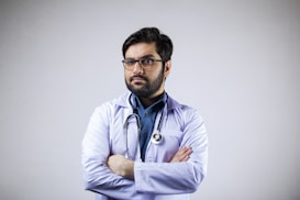 A man with a beard and glasses is wearing a white coat and a stethoscope around his neck. His arms are crossed, and he appears confident and professional. The background is plain and light-colored.