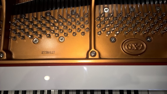 The image displays the interior mechanics of a piano, highlighting the shiny metal pins, strings, and tuning pegs against the gold-colored frame. The serial number 2739127 and the model marking 'GX-2' are visible on the frame. Below, a portion of the keyboard is partially visible, with white and black keys aligned in parallel.