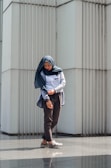 A person in casual attire stands in front of a modern, textured wall made of vertical panels. The person is wearing a hijab and looking downwards with a contemplative expression, hands slightly adjusted at the sleeves. The sunlight casts a gentle shadow on the ground, creating a reflective surface.