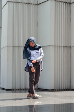 A person in casual attire stands in front of a modern, textured wall made of vertical panels. The person is wearing a hijab and looking downwards with a contemplative expression, hands slightly adjusted at the sleeves. The sunlight casts a gentle shadow on the ground, creating a reflective surface.