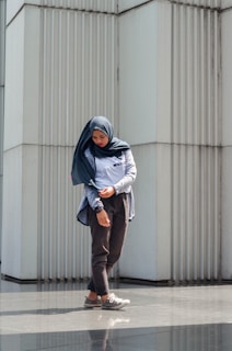 A person in casual attire stands in front of a modern, textured wall made of vertical panels. The person is wearing a hijab and looking downwards with a contemplative expression, hands slightly adjusted at the sleeves. The sunlight casts a gentle shadow on the ground, creating a reflective surface.