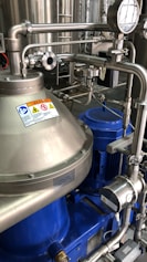 A complex assembly of stainless steel industrial machinery with pipes and valves, featuring a control gauge and warning signs. The machinery components are metallic with blue accents, indicating a possible connection to liquid processing or fluid control.