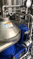 A complex assembly of stainless steel industrial machinery with pipes and valves, featuring a control gauge and warning signs. The machinery components are metallic with blue accents, indicating a possible connection to liquid processing or fluid control.