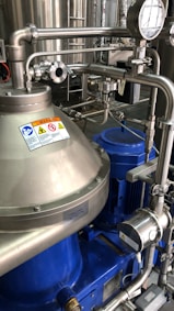 A complex assembly of stainless steel industrial machinery with pipes and valves, featuring a control gauge and warning signs. The machinery components are metallic with blue accents, indicating a possible connection to liquid processing or fluid control.