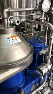 A complex assembly of stainless steel industrial machinery with pipes and valves, featuring a control gauge and warning signs. The machinery components are metallic with blue accents, indicating a possible connection to liquid processing or fluid control.