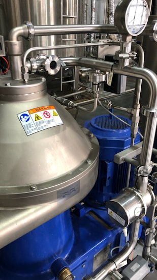 A complex assembly of stainless steel industrial machinery with pipes and valves, featuring a control gauge and warning signs. The machinery components are metallic with blue accents, indicating a possible connection to liquid processing or fluid control.