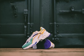 A pair of colorful sneakers is placed on the ground in front of a large, dark metal door. The sneakers feature a mix of colors including teal, yellow, white, and purple, with a prominent logo on the side. The setting gives a slightly urban feel with the focus on the vibrant shoes against the muted background.
