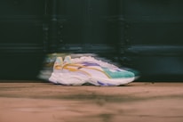 A pair of stylish sneakers with a colorful design, including shades of green, yellow, purple, and white. The sneakers are positioned on a wooden surface with a blurred background, creating a sense of motion.