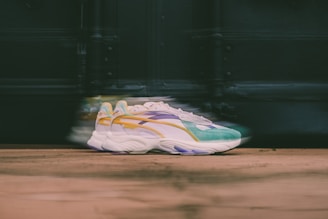 A pair of stylish sneakers with a colorful design, including shades of green, yellow, purple, and white. The sneakers are positioned on a wooden surface with a blurred background, creating a sense of motion.