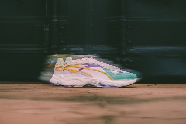 A pair of stylish sneakers with a colorful design, including shades of green, yellow, purple, and white. The sneakers are positioned on a wooden surface with a blurred background, creating a sense of motion.