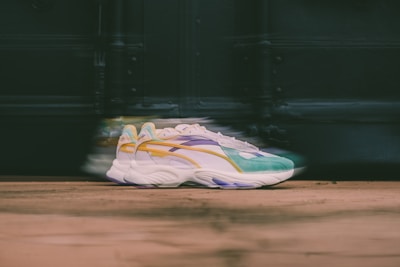 A pair of stylish sneakers with a colorful design, including shades of green, yellow, purple, and white. The sneakers are positioned on a wooden surface with a blurred background, creating a sense of motion.