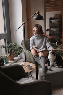 woman in gray sweater sitting on chair beside black and white cat