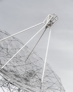A large metal satellite dish with a mesh design, angled against a pale sky. The framework consists of white poles and intricate wire netting that creates geometric patterns.