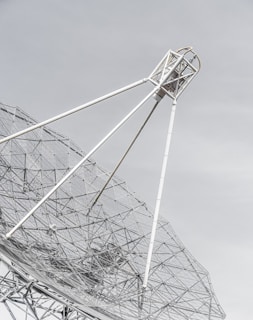 A large metal satellite dish with a mesh design, angled against a pale sky. The framework consists of white poles and intricate wire netting that creates geometric patterns.