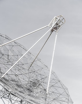 A large metal satellite dish with a mesh design, angled against a pale sky. The framework consists of white poles and intricate wire netting that creates geometric patterns.