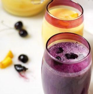 Two glasses filled with smoothies, one purple with visible berries on top and another yellow with a chunk of fruit. Several cherries and orange slices are scattered on a white surface nearby.