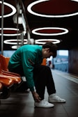 A person is seated in a modern, dimly lit waiting area with circular overhead lights and vivid orange seating. The individual is wearing a teal jacket and white sneakers, appearing to tie their shoelaces. The floor is tiled, and the space has a sleek, industrial design with a somewhat moody ambiance.