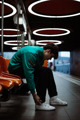 A person is seated in a modern, dimly lit waiting area with circular overhead lights and vivid orange seating. The individual is wearing a teal jacket and white sneakers, appearing to tie their shoelaces. The floor is tiled, and the space has a sleek, industrial design with a somewhat moody ambiance.