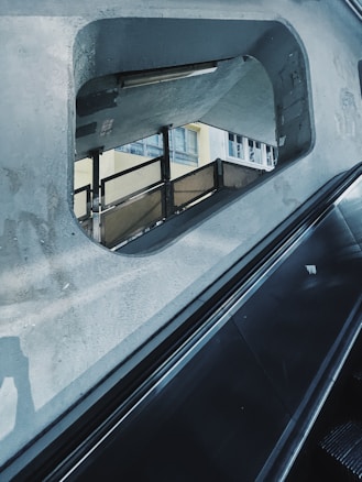 A viewing window in a concrete structure reveals a scene with an interior stairwell and windows of a building. An escalator rail runs diagonally across the foreground, showing reflections on its metallic surface. The concrete has a worn texture.