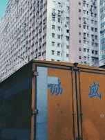 A large residential building with numerous windows is visible, showcasing a densely packed urban environment. The building has a faded, slightly worn appearance. In front of it, there is a brightly colored orange structure, possibly a truck or storage container, featuring blue Chinese characters.