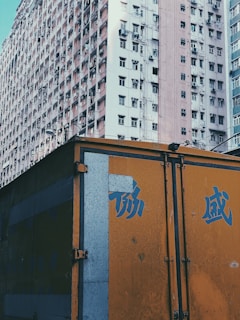 A large residential building with numerous windows is visible, showcasing a densely packed urban environment. The building has a faded, slightly worn appearance. In front of it, there is a brightly colored orange structure, possibly a truck or storage container, featuring blue Chinese characters.