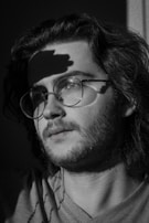 A person with long hair and glasses is captured in a black and white portrait. The lighting creates a shadow across the person's face, adding a dramatic effect. The individual looks away from the camera, contributing to a contemplative mood.