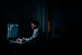 A person is sitting at a desk in a dimly lit room, wearing headphones and looking intently at a large computer screen with dark-themed code editor or software visible. The focus suggests concentration, and there is a mug or cup on the desk, indicating a prolonged working session.