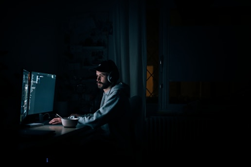 A focused portrait of Arif Hossen working intently at his computer in a dimly lit room with code visible on the screen.
