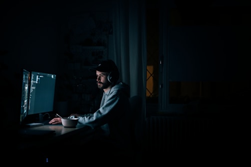 A person is sitting at a desk in a dimly lit room, wearing headphones and looking intently at a large computer screen with dark-themed code editor or software visible. The focus suggests concentration, and there is a mug or cup on the desk, indicating a prolonged working session.