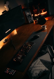 A workspace setup featuring a large monitor with the words 'CREATE MORE', a mechanical keyboard with backlighting, a gaming mouse, a desktop computer with visible internal lighting, a microphone, and a speaker. A mug sits on the desk beside a remote control and a pillow lies on the floor.