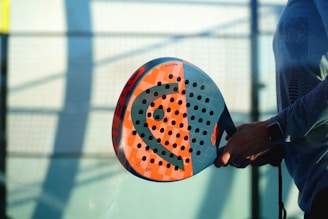 A person holding a colorful padel racket with a perforated design, standing against a blurred background of a sports court. The lighting creates shadows, adding depth to the scene.