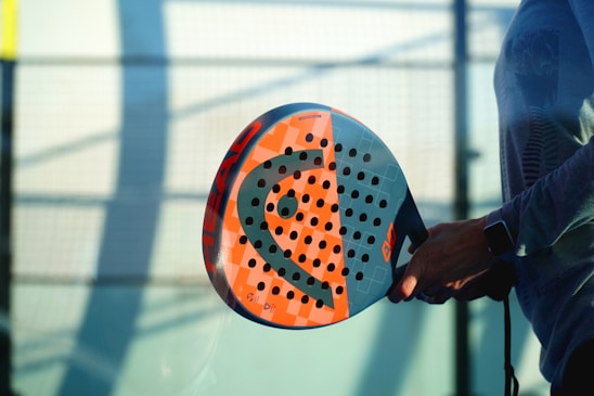 A person holding a colorful padel racket with a perforated design, standing against a blurred background of a sports court. The lighting creates shadows, adding depth to the scene.