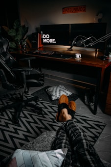 A cozy room setup features a desk with dual monitors displaying the words 'Do More'. The desk also holds a microphone, speakers, a cup, and a keyboard. A gaming chair is positioned nearby. On the floor, a patterned rug, pillows, and slippers with feet in them can be seen. The ambiance is warm and relaxed.
