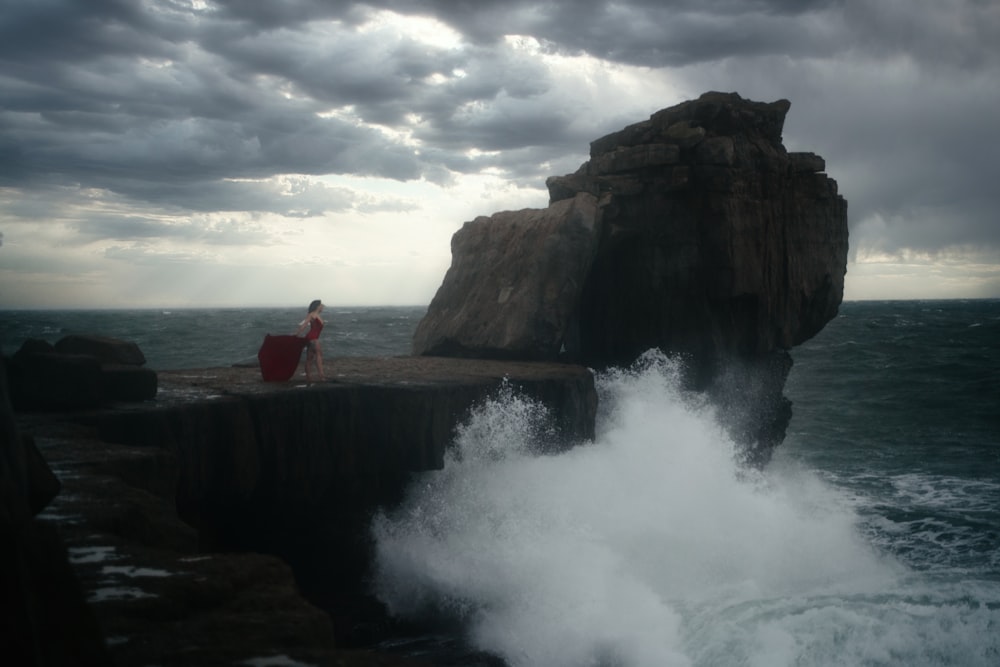 man in red shirt standing on rock formation near sea water during daytime