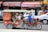A motorbike is pulling a tuk-tuk-style carriage with several passengers inside. The driver wears a helmet and a blue shirt, while the passengers include children and an adult, with red curtains partially drawn on the carriage. The background captures motion blur, suggesting movement, and there's a visible bustling street scene with various vehicles and signage.