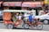 A motorbike is pulling a tuk-tuk-style carriage with several passengers inside. The driver wears a helmet and a blue shirt, while the passengers include children and an adult, with red curtains partially drawn on the carriage. The background captures motion blur, suggesting movement, and there's a visible bustling street scene with various vehicles and signage.