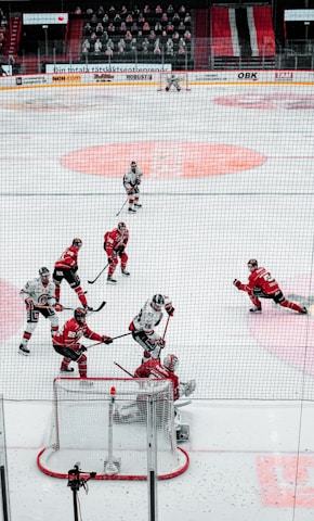 A group of ice hockey players are in the middle of a game on the ice rink. Some players are wearing red uniforms while others are in white, engaging in an intense moment near the goal. The goalie in red is positioned to block a shot from the opposing team. The audience is visible in the background, seated in the stands.