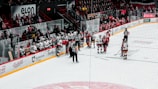 A group of ice hockey players gather on the rink near the benches. Players from both teams, wearing red and white jerseys, are engaged in discussion. Referees in black and white striped uniforms stand nearby. A row of seated players and coaches are on the benches with a crowd in the background.