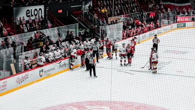A group of ice hockey players gather on the rink near the benches. Players from both teams, wearing red and white jerseys, are engaged in discussion. Referees in black and white striped uniforms stand nearby. A row of seated players and coaches are on the benches with a crowd in the background.