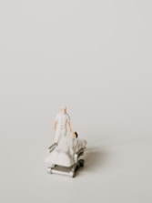 A miniature figurine of a nurse stands beside a hospital bed with a patient lying under a white blanket. The scene is minimalist with a pale background, emphasizing the medical figures.
