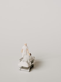 A miniature figurine of a nurse stands beside a hospital bed with a patient lying under a white blanket. The scene is minimalist with a pale background, emphasizing the medical figures.