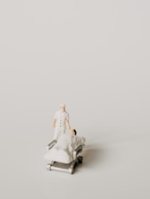 A miniature figurine of a nurse stands beside a hospital bed with a patient lying under a white blanket. The scene is minimalist with a pale background, emphasizing the medical figures.
