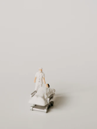 A miniature figurine of a nurse stands beside a hospital bed with a patient lying under a white blanket. The scene is minimalist with a pale background, emphasizing the medical figures.