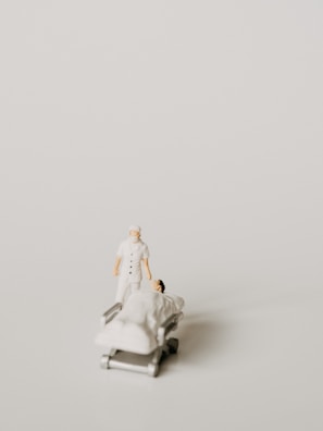 A miniature figurine of a nurse stands beside a hospital bed with a patient lying under a white blanket. The scene is minimalist with a pale background, emphasizing the medical figures.
