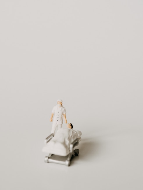 A miniature figurine of a nurse stands beside a hospital bed with a patient lying under a white blanket. The scene is minimalist with a pale background, emphasizing the medical figures.