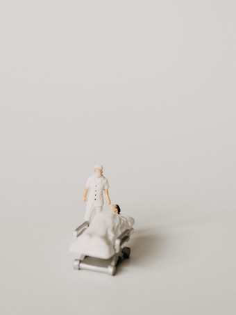 A miniature figurine of a nurse stands beside a hospital bed with a patient lying under a white blanket. The scene is minimalist with a pale background, emphasizing the medical figures.