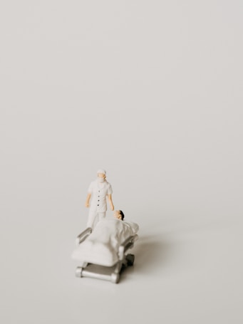 A miniature figurine of a nurse stands beside a hospital bed with a patient lying under a white blanket. The scene is minimalist with a pale background, emphasizing the medical figures.