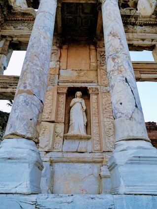 A classical statue is positioned between two large, ornate marble columns. The statue, which depicts a robed figure, is set within an intricately carved alcove on an ancient stone structure. The surface of the columns and the surrounding stonework showcase visible wear and cracks, indicating the heritage and age of the architecture.
