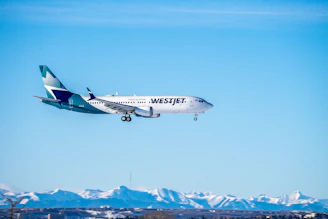A sleek airplane soaring above snow-capped mountains under a bright teal sky.