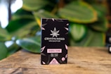 A black box of Crystalweed CBD product is placed on a wooden surface with a blurred background of green leaves. The packaging features a cannabis leaf design and text in pink and white, highlighting the product's flavor as bubble gum and its natural ingredients.